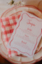 A wedding menu with a whimsy pink font, wavy pink edges and pale pink background stripes on white card. Placed on cream plates with pink edges detailing and a red gingham napkin.