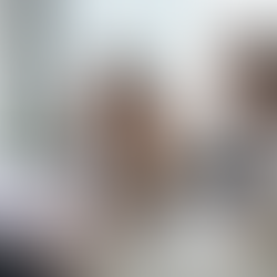 Professional meeting between three colleagues at a table, with one man gesturing while holding a pen, a woman listening thoughtfully with her hand to her head, and another man working on a laptop. Papers with charts and a coffee cup are on the table in a bright office setting.