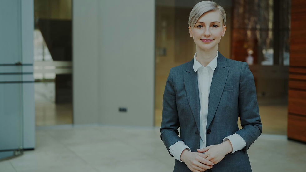 Professional woman with short blonde hair wearing a grey business suit and white shirt, standing in a modern office environment and smiling at the camera.