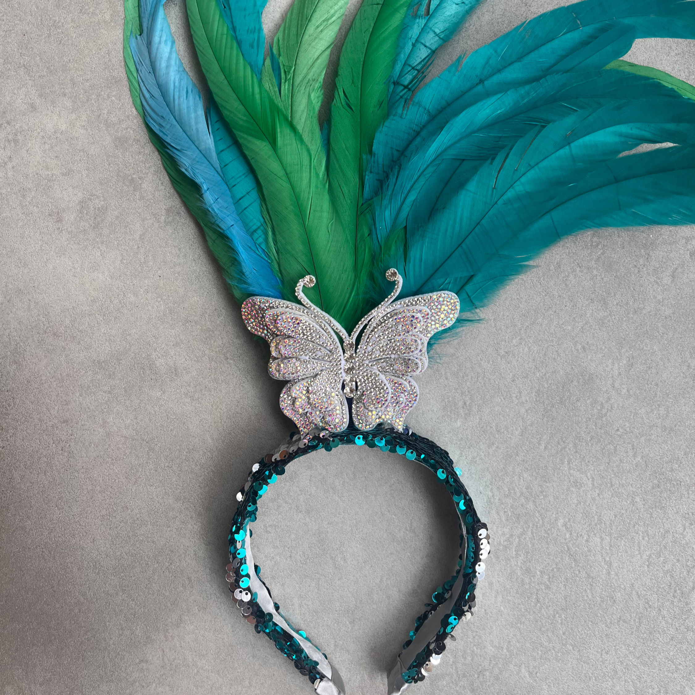 A tiara é revestida de lantejoulas azuis e prateadas com uma borboleta de strass no topo e penas verde-água e turquesa atrás.