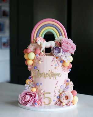 Pretty pink buttercream cake with a unicorn figure, fondant pastel rainbow, baked doughnuts, and artificial flowers