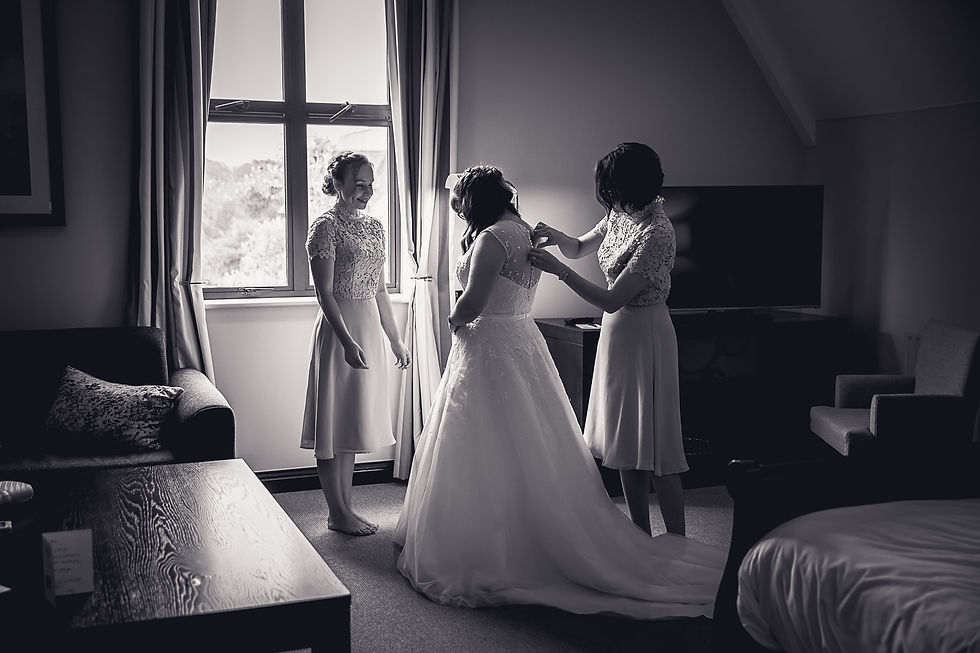 A bride and her bridesmaid stand in a hotel room. One of the bridesmaid is doing up the bride's dress
