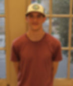 A young white man stands in front of a window, wearing a rust-colored t-shirt and a baseball cap.