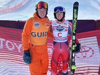 Two skiers in bright gear smile at the camera. One wears orange with "GUIDE," the other in red with a ski. Snowy mountain background.