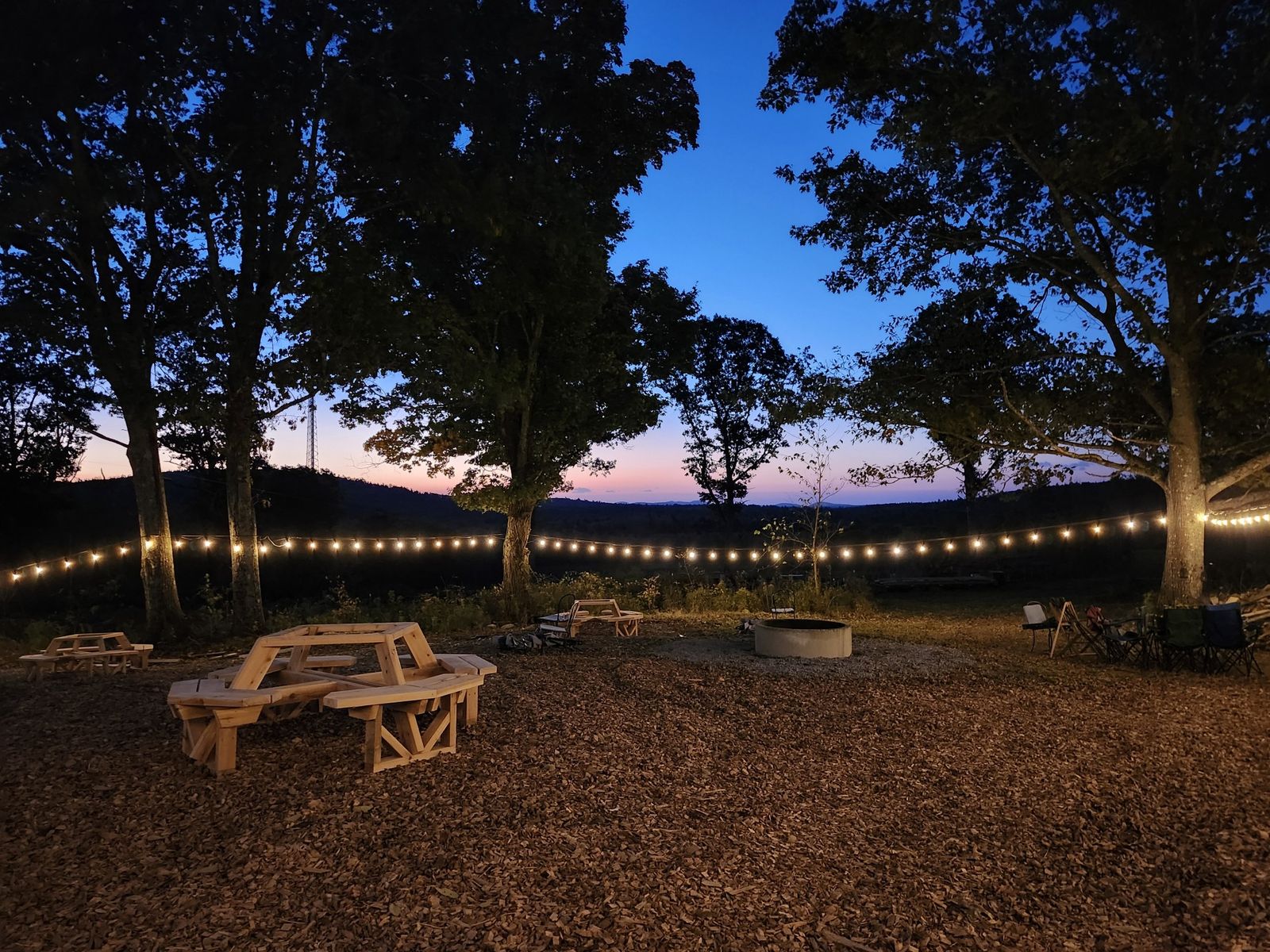 The outdoor seating area at dusk, with picnic tables and a fire pit under string lights and a view of the hills