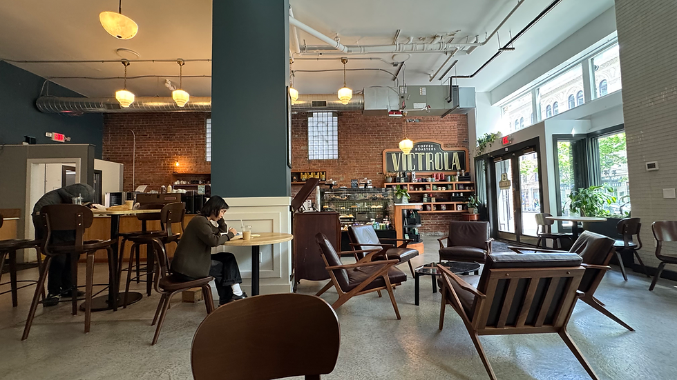 Coffee shop with brick walls, wooden chairs, and a "Victrola" sign. Two people sit with drinks, one studying. A cozy, relaxed setting.