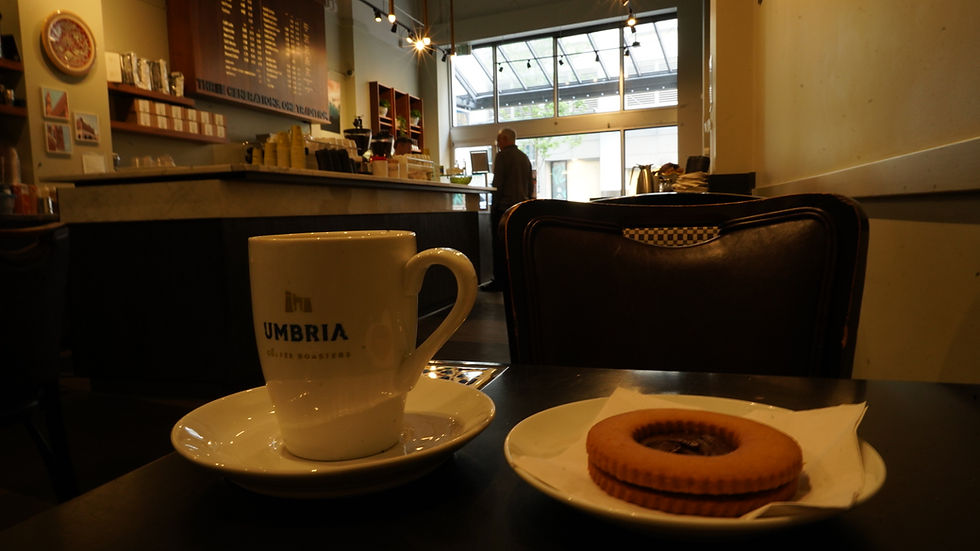 Coffee cup with "Umbria" logo and a cookie on a table in a cozy café. Warm lighting, menu board, and a person at the counter in the background.