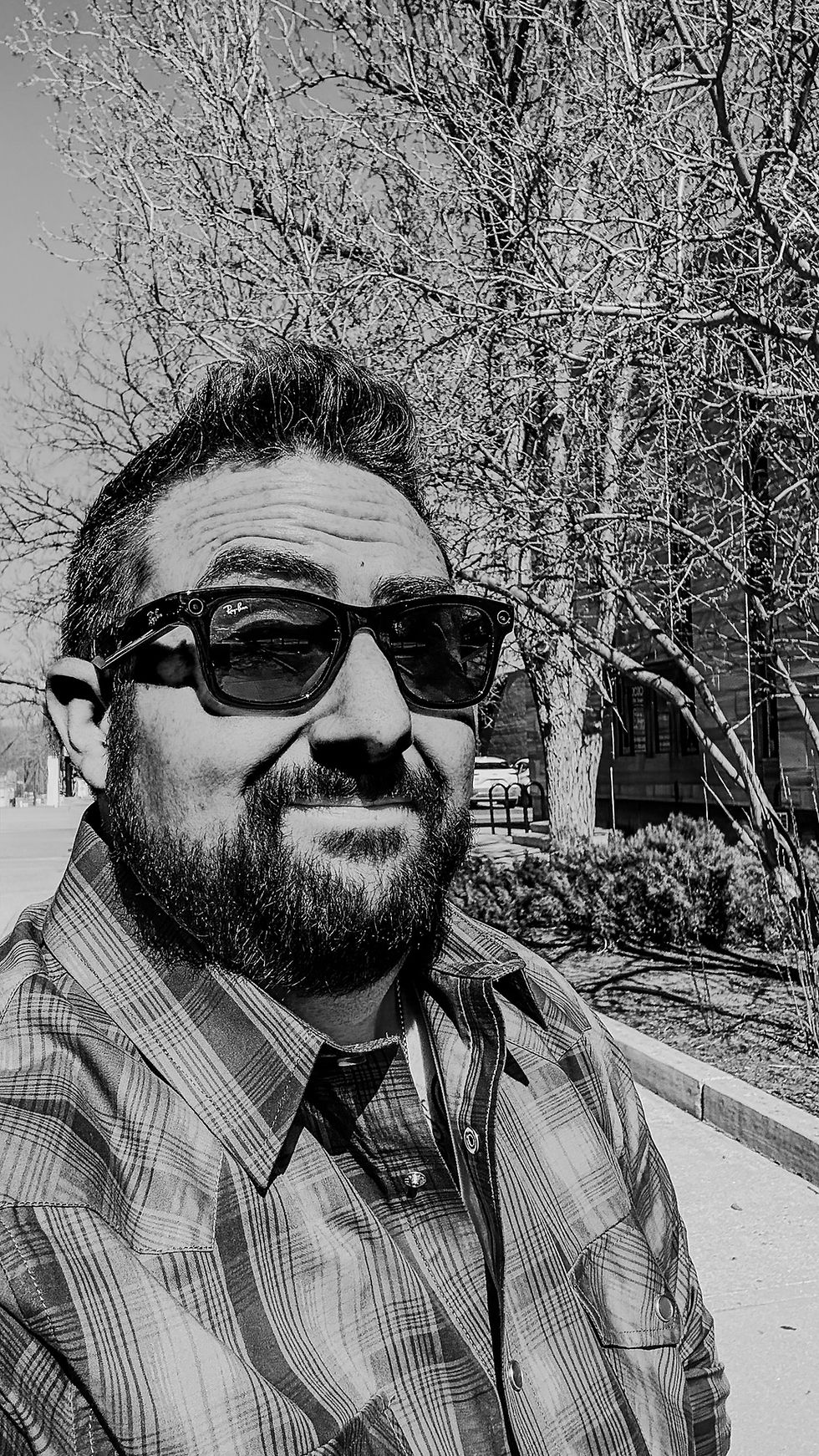 Bearded man wearing sunglasses and a plaid shirt outdoors.