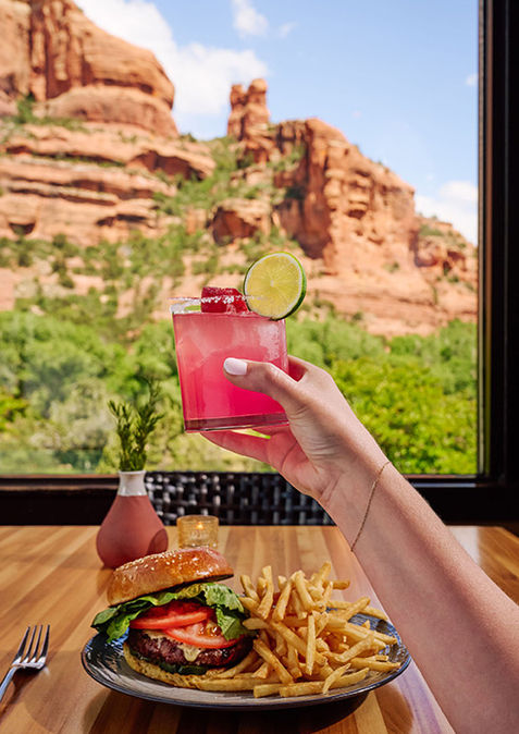 A hand holds a pink cocktail with a lime slice in front of a window view of red rock formations. A plate with a burger and fries is on a wooden table.