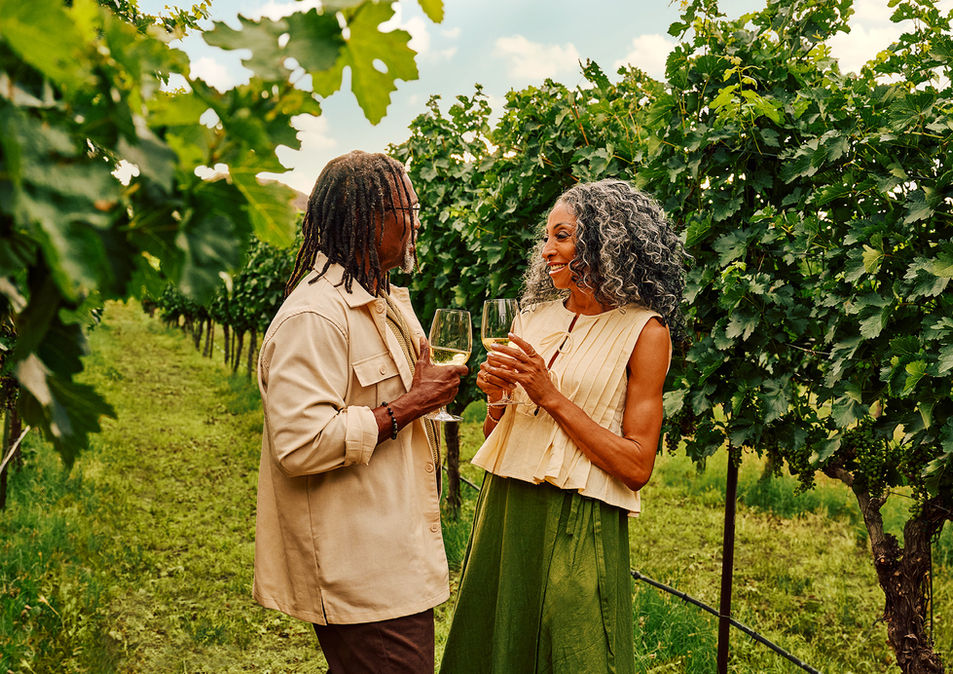 A joyful couple stands in a lush vineyard, holding glasses of white wine. They smile warmly at each other, surrounded by vibrant green grapevines under a clear sky.