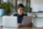 A female auditor sits at her desk, examining some physical paperwork while her laptop is open.