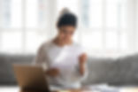 A woman of Indian descent sits in her living room near her laptop, reading a stack of paperwork