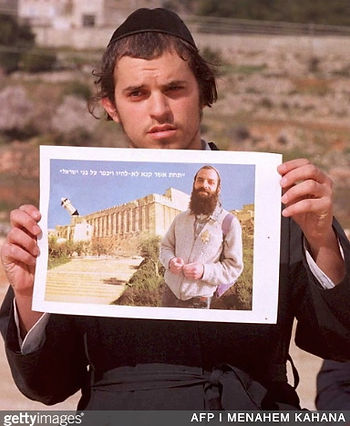 An orthodox Jew holds a photo of Baruch Goldstein. The photo has been altered to show Goldstein standing in front of the Tomb of the Patriarchs, the photo includes a destroyed minaret.