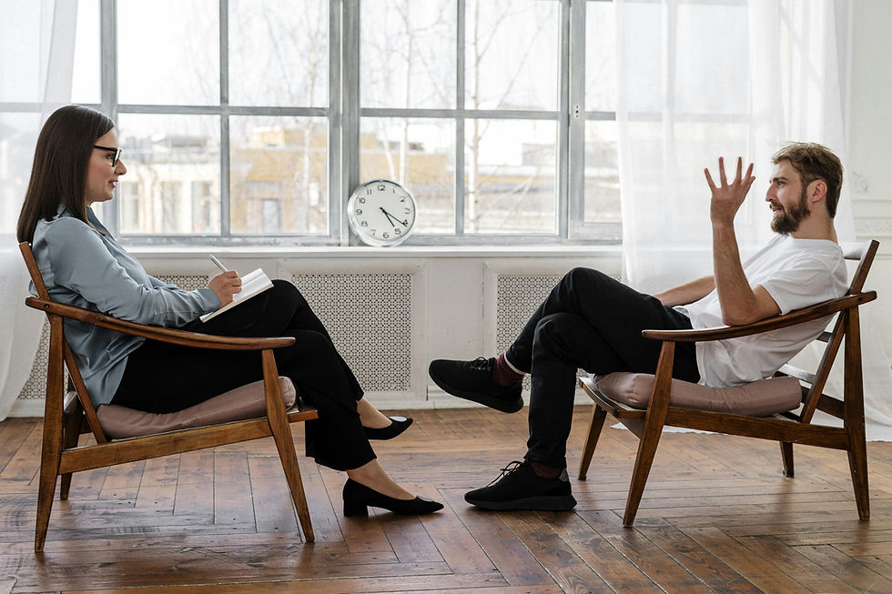 A man and woman converse in chairs by a window. She writes notes while he gestures. A clock shows 12:10. Room is well-lit with wooden floor.