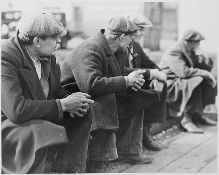 Row of men sitting down all wearing trench coats and flat caps.