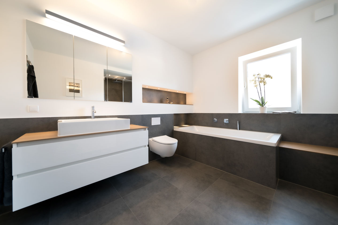 Modernes WC-Design: Wandhängendes WC vor dunkler Rückwand mit integriertem Echtholz-Regal für Handtücher – elegante Stauraumlösung von BAD ELEMENTE Hamburg.