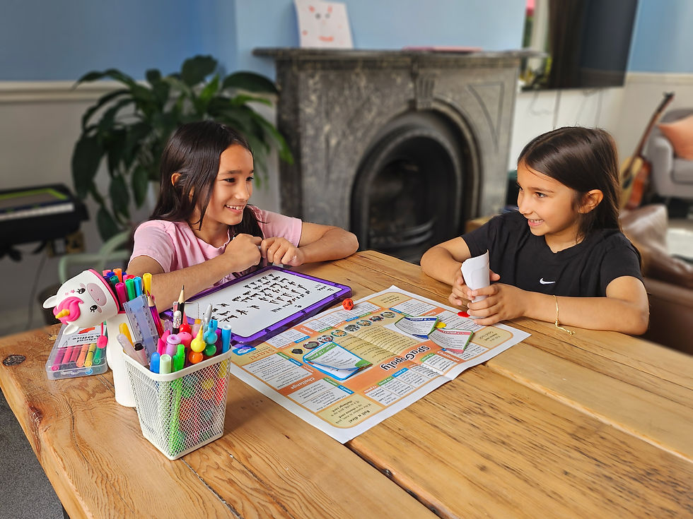 Two girls with brown hair sat at a wooden table in a living room. On the table is a pot of colourful pens, a whiteboard and a school book.