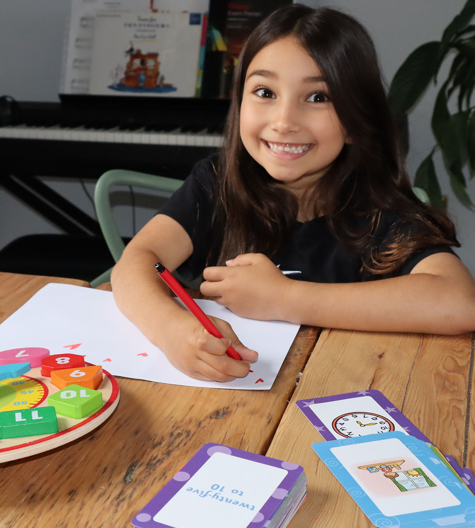 A young girl with brown hair smiling, using a red pen to draw hearts on a white peice of paper. On the table in front of her is a set of educational cards used to teach how to tell the time.