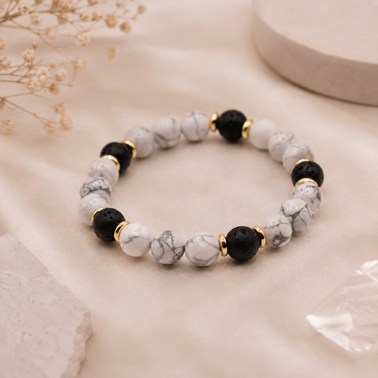 Howlith Edelsteinarmband mit Lavastein und goldfarbenen Akzenten auf edlem Stoffhintergrund.