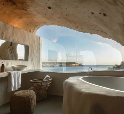 A luxurious, cave-inspired bathroom interior featuring organic, sculpted beige walls and a minimalist stone bathtub. A large, frameless panoramic window dominates the room, offering a breathtaking view of a blue ocean and distant limestone cliffs. Sustainable design elements include textured earthen plaster, a floating vanity with a vessel sink, and woven wicker seating, all bathed in soft natural light.