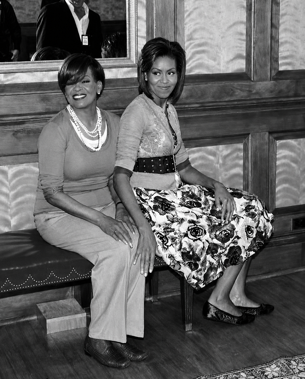 Two smiling women sit on a bench in a wood-paneled room, one in a floral skirt and pearls, with a mirror behind them.