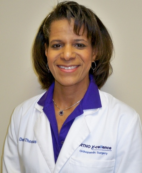 Smiling person in white lab coat with blue shirt. Coat reads Ortho X-celence Orthopaedic Surgery. Neutral background.