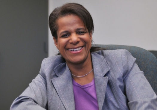 Smiling woman in a purple shirt and gray blazer sits in an office chair. Gray and white background, creating a friendly and professional mood.