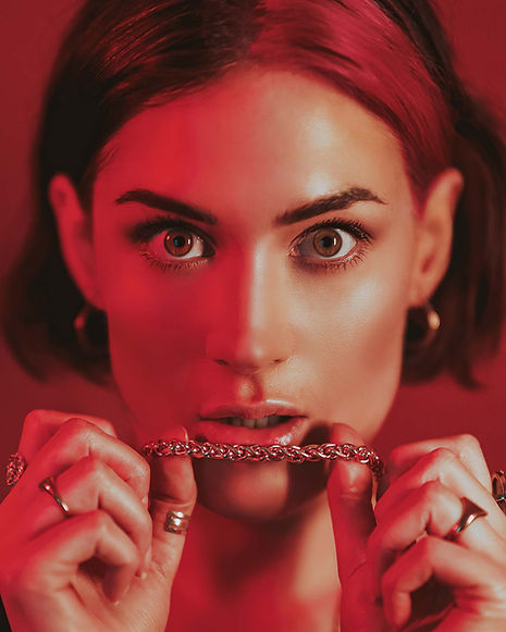 Face on portrait of a woman lit with red lighting, holding a gold necklace against their open mouth.