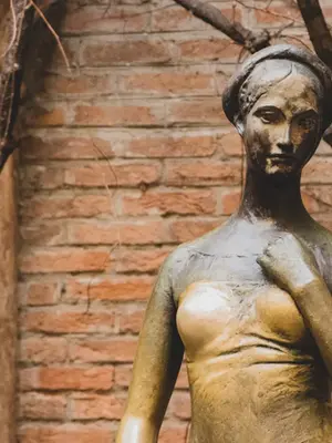 Um plano médio aproximado da famosa estátua de bronze de Julieta Capuleto na Casa di Giulietta, em Verona. A escultura apresenta uma jovem com expressão serena e melancólica, com o cabelo preso sob uma tiara delicada. O bronze exibe uma pátina escura e esverdeada pelo tempo, exceto na região do seio direito, que brilha com um tom dourado polido devido ao toque frequente de visitantes em busca de sorte no amor. Ao fundo, uma parede rústica de tijolos aparentes em tons de terracota é emoldurada por galhos secos e retorcidos de uma trepadeira, criando uma atmosfera romântica e histórica.