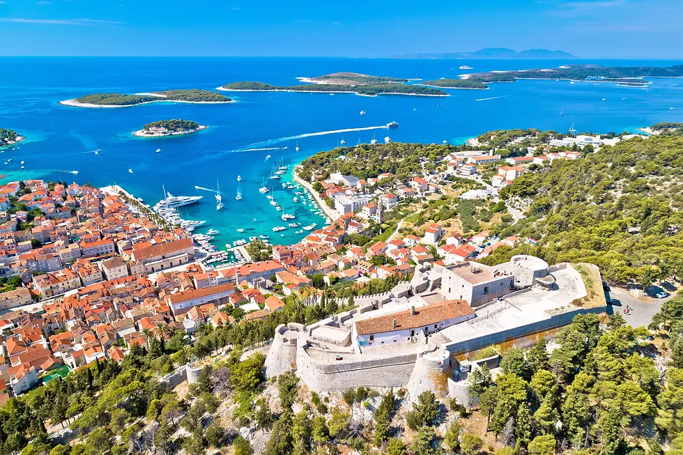 Vista aérea panorâmica de Hvar sob um céu azul profundo e ensolarado. Em primeiro plano, destaca-se a imponente Fortaleza de Hvar (Fortica), construída em pedra clara com muralhas robustas e baluartes circulares que dominam a colina. Abaixo da fortaleza, a encosta verdejante desce até à cidade costeira, caracterizada por um mar denso de telhados de terracota laranja. O porto em forma de ferradura está repleto de iates brancos e barcos de recreio em águas azul-turquesa cristalinas. Ao fundo, o arquipélago das Ilhas Pakleni espalha-se pelo mar Adriático, completando o cenário paradisíaco.