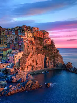 Vista panorâmica de Manarola ao pôr do sol. A aldeia é composta por casas coloridas em tons de terracota, amarelo e rosa, empilhadas num penhasco íngreme sobre o Mar da Ligúria. À esquerda, barcos de pesca azuis e brancos estão estacionados num caminho de pedra junto à água. O céu apresenta um gradiente de azul e rosa vibrante, cujas cores se refletem suavemente na superfície calma do mar. Rochas escuras emergem da água junto à costa, e uma pequena doca de pedra estende-se em direção ao mar à direita.