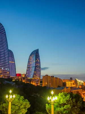Uma fotografia panorâmica da paisagem urbana de Baku, no Azerbaijão, capturada durante o crepúsculo. O ponto focal são as três Flame Towers, arranha-céus em formato de chama com fachadas de vidro iluminadas por linhas horizontais de LED em tons de azul e roxo. À esquerda, uma encosta arborizada esconde parte da base das torres, onde se nota a silhueta de uma mesquita iluminada. Em primeiro plano, dois postes de luz ornamentais com globos brilhantes emolduram a vista. A cidade ao fundo exibe uma malha densa de edifícios com luzes douradas, estendendo-se até o horizonte sob um céu que transita do azul profundo para um tom suave de laranja e amarelo.