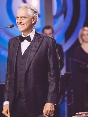 Fotografia de plano médio do tenor Andrea Bocelli a cantar com os olhos fechados e um sorriso sereno num palco iluminado. Ele veste um fato de gala preto com laço; ao fundo, de forma desfocada, veem-se músicos de uma orquestra, uma harpa e elementos de iluminação cénica em tons de azul e roxo.