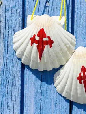 Um detalhe em close-up de duas conchas de vieira brancas, símbolo tradicional dos peregrinos do Caminho de Santiago, penduradas por cordões amarelos vibrantes. Cada concha apresenta a Cruz de Santiago pintada em vermelho vivo no centro. Elas estão posicionadas contra uma parede de madeira rústica pintada de azul claro, cujas tábuas verticais mostram texturas de desgaste e descascamento, criando um contraste visual marcante entre o branco das conchas, o vermelho da cruz e o fundo azul.