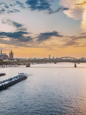 Uma ampla fotografia panorâmica do Rio Reno em Colónia, Alemanha, sob um céu azul límpido. No centro, uma grande barcaça de carga navega pelas águas calmas do rio, deixando um rasto de espuma branca. À direita, a silhueta monumental da Catedral de Colónia, com as suas duas torres gémeas góticas, domina o horizonte da cidade. Ao lado da catedral, avistam-se edifícios coloridos e a Ponte Hohenzollern ao longe. As margens do rio são ladeadas por árvores verdes e caminhos de passeio. A iluminação solar brilhante destaca a serenidade da paisagem fluvial e a grandiosidade da arquitetura histórica e moderna da cidade.