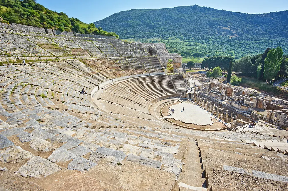 Uma fotografia panorâmica de ângulo elevado capturando o Grande Teatro de Éfeso, na Turquia. A imagem mostra a vasta arquibancada semicircular (cavea) de pedra clara e desgastada que sobe a encosta da montanha. No centro inferior, o palco plano (orquestra) é visível com alguns turistas caminhando sobre ele. À direita do palco, restam as ruínas da fachada da cena (skene), com fileiras de colunas de mármore e blocos de pedra caídos. O plano de fundo exibe uma montanha verdejante sob um céu azul limpo. A iluminação é natural e direta do sol, realçando o relevo dos degraus de pedra e as texturas das ruínas clássicas.