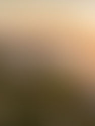 Uma fotografia aérea panorâmica capturando o Cabo de Finisterre na Galiza, Espanha, durante o pôr do sol. No centro da imagem, destaca-se o complexo do Farol de Finisterre construído sobre um promontório rochoso escarpado que se projeta em direção ao mar. O farol possui uma torre de pedra marrom cilíndrica rodeada por edifícios anexos caiados de branco e com telhados terracota. Outras pequenas casas tradicionais estão espalhadas pela encosta gramada e rochosa. À direita, o sol está se pondo no horizonte sobre o oceano, banhando toda a cena com uma luz dourada e quente que se reflete na superfície da água. O céu apresenta tons suaves de amarelo, laranja e azul claro. Uma pequena estrada sinuosa corta a vegetação verde do penhasco à esquerda.