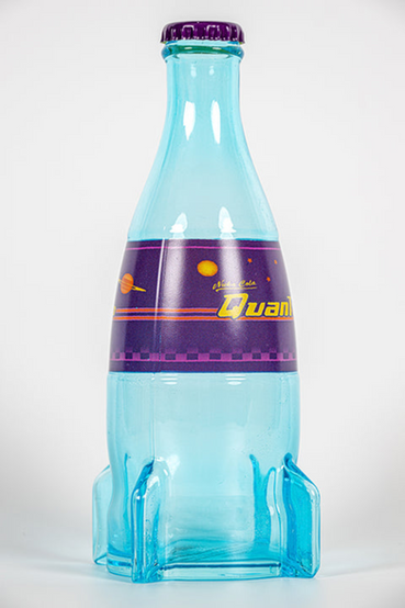 A glass replica of Nuka-Cola Quantum from Fallout soft drink in the rocket bottle design as seen in Fallout 4 and in Fallout 76, the bottle itself is tinted light blue, with the soda label saying 'Nuka-Cola Quantum' in yellow text on a purple background. Also pictured are loose metal bottlecaps that match the Nuka-Cola Quantum soda label
