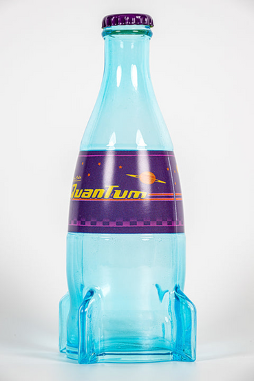 A glass replica of Nuka-Cola Quantum from Fallout soft drink in the rocket bottle design as seen in Fallout 4 and in Fallout 76, the bottle itself is tinted light blue, with the soda label saying 'Nuka-Cola Quantum' in yellow text on a purple background. Also pictured are loose metal bottlecaps that match the Nuka-Cola Quantum soda label