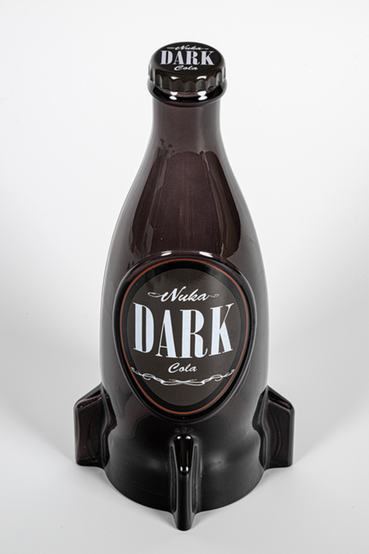 A glass replica of Nuka-Cola Dark (Nuka Dark) from Fallout soft drink in the rocket bottle design. The bottle itself is tinted in a blackish brown color, with front soda label saying 'Nuka Dark Cola' in white on a dark brown background. Also pictured are loose metal bottlecaps that match the Nuka-Cola Dark label