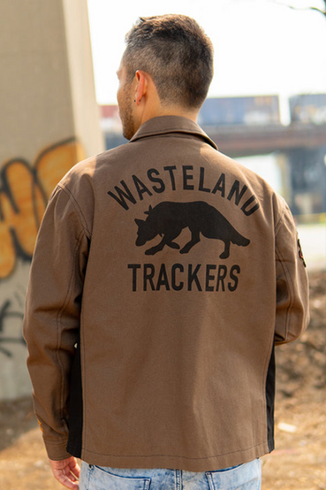 Dogmeat from Fallout 4 inspired canvas jacket in brown with dark brown accents. It also features red paisley lining, Dogmeat's silhouette with 'Wasteland Trackers' printed on the back, DOGMEAT embroidered above the front left pocket, Dogmeat emoji patch on the right sleeve, and embroidered crossbones on top of the left sleeve cuff