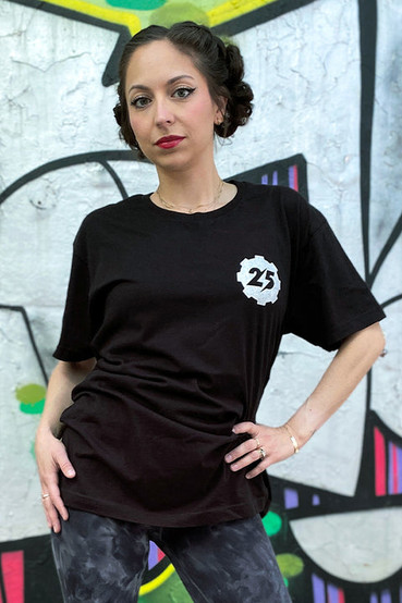 Black graphic t-shirt as worn by a male and female model, t-shirt has a small gear shape on the front left side, and a much larger one on the back. The gear logo has the number 25 in the middle, the negative space between the numbers is Fallout's lighting bolt symbol.
