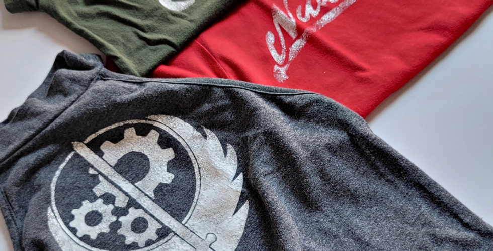 Brotherhood of Steel gear logo from Fallout in white stenciled on a dark grey crop top, Reilly's Rangers clover logo from Fallout in white stenciled on an olive green shirt, and Nuka-Cola script logo from Fallout in white stenciled on a red shirt