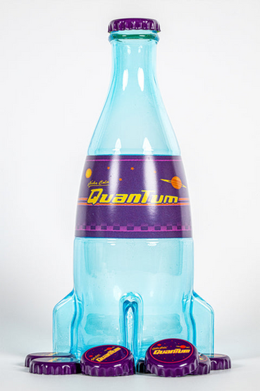 A glass replica of Nuka-Cola Quantum from Fallout soft drink in the rocket bottle design as seen in Fallout 4 and in Fallout 76, the bottle itself is tinted light blue, with the soda label saying 'Nuka-Cola Quantum' in yellow text on a purple background. Also pictured are loose metal bottlecaps that match the Nuka-Cola Quantum soda label