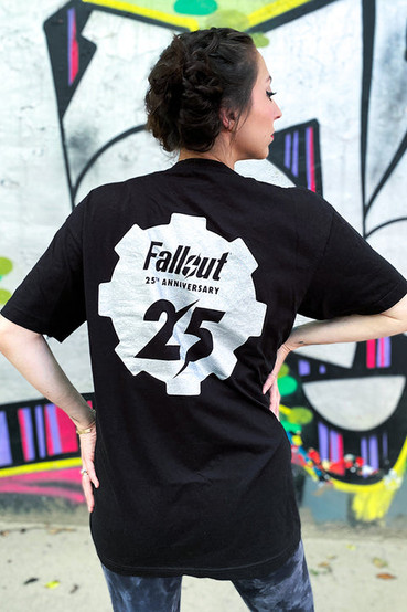 Black graphic t-shirt as worn by a male and female model, t-shirt has a small gear shape on the front left side, and a much larger one on the back. The gear logo has the number 25 in the middle, the negative space between the numbers is Fallout's lighting bolt symbol.