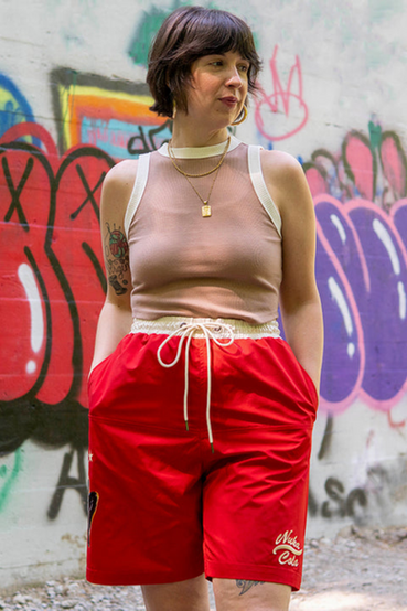Nuka Girl from Fallout themed shorts in red, features Nuka Girl doing a classic pin-up pose with her Nuka Gun printed on the front right side, and 3D embroidered Nuka-Cola text logo in cream-colored thread on the front bottom left
