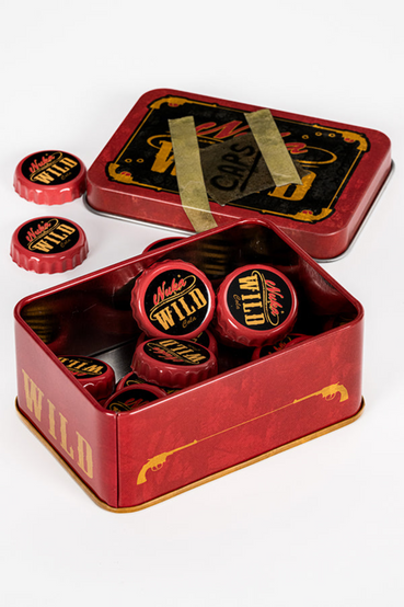 Nuka-Cola Wild from Fallout themed metal tin that comes with 20 matching bottle caps. The front of the tin has a Nuka-Cola Wild label that has the look of a handwritten note that says 'CAPS' taped on front, all gold, black and red colorway. The loose bottle caps have the Nuka-Cola Wild logo in a cowboy script font on the front of the cap, with the inside of the bottle cap a silver color