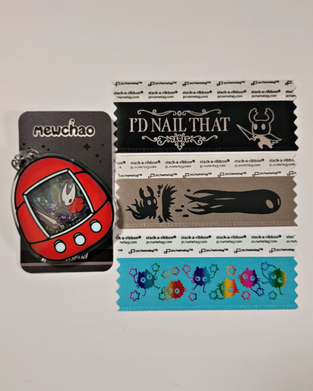 Hornet from Hollow Knight as a Tamagochi inspired acrylic shaker on the left, Hollow Knight and cat soot sprite badge ribbons on the right
