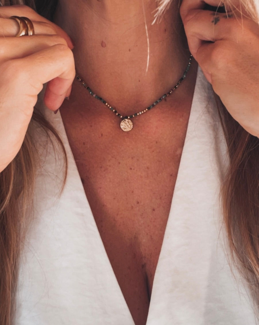 Un collier délicat composé de perles multicolores et d’une médaille dorée texturée. Les nuances vertes et roses s’accordent p