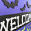 Thumbnail: A round, wooden purple and black Halloween welcome sign with white letters shaped like bones and decorative lime green bats.